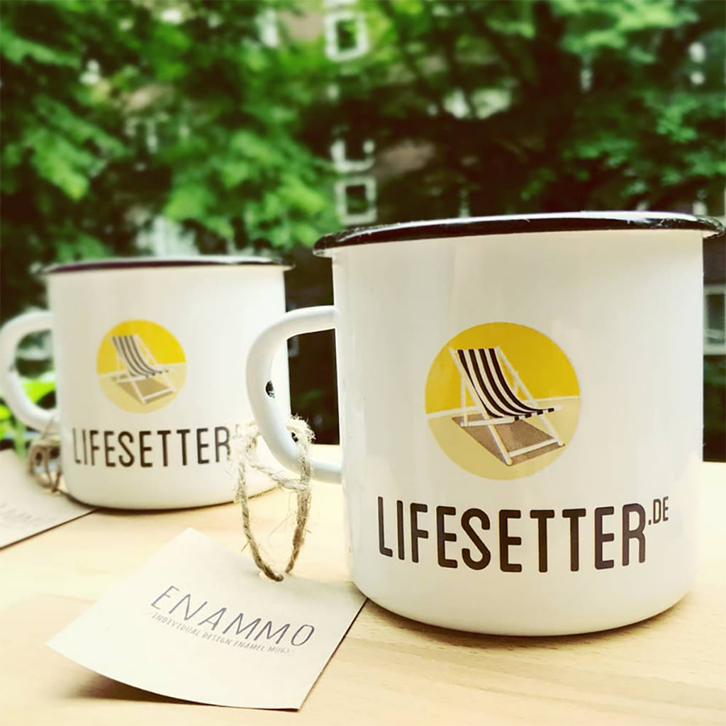 Two Lifesetter enamel mugs on a table with a blurred green outdoor background