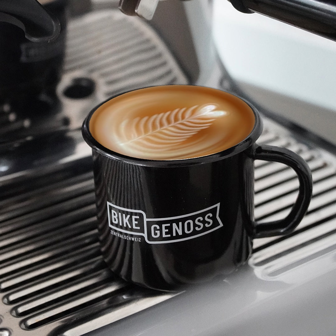 Black custom printed enamel mug with 'BIKE GENOSS' branding holding a cappuccino in front of an espresso machine.