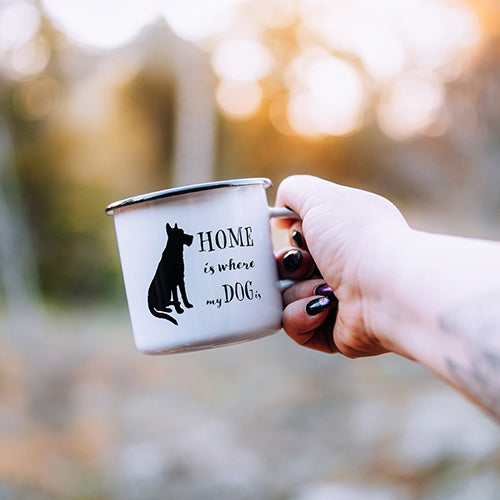 Person holding enamel mug with 'Home is where my dog is' text and dog silhouette against a blurred natural background