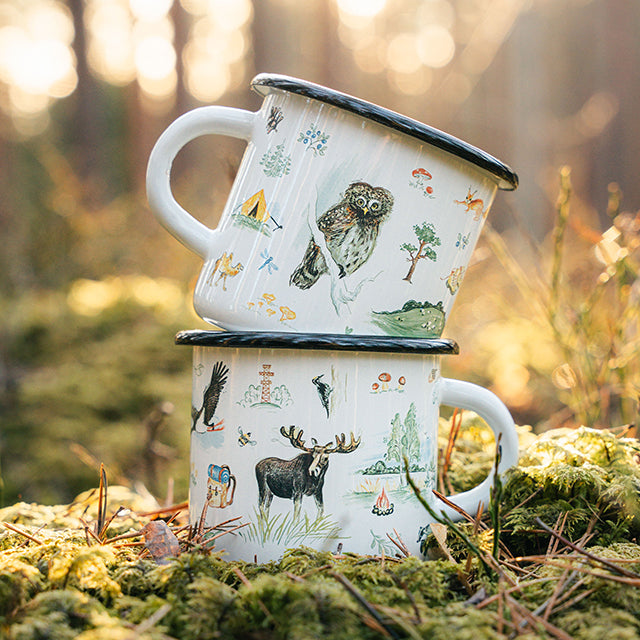 Two custom printed enamel mugs with nature-themed illustrations stacked on a mossy surface in a forest setting